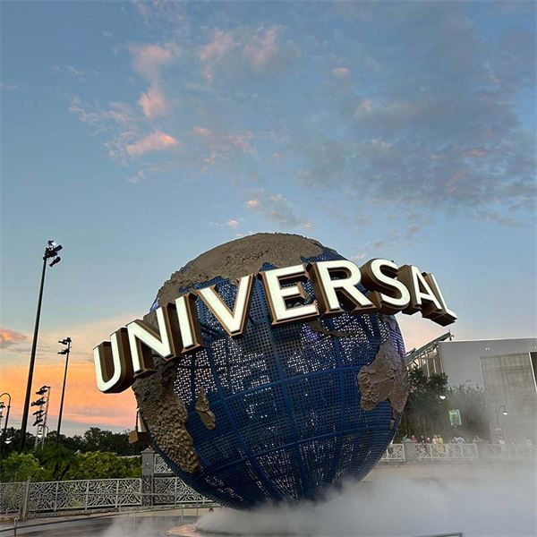 The revolving Universal globe at dusk, lit gold against a pink and orange sky with mist drifting around its base.